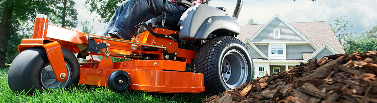 An orange riding mower being driven though a yard next to a small pile of bark chips